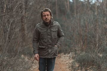 Blond man in green coat walks on a pathway between trees in nature reserve.