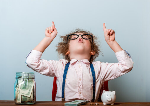 Funny little kid girl in glasses with fingers pointing up. Child boss holding dollars in jar. thumb up to the side smiling happy with open