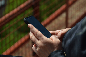 Senior hands holding a smartphone with a metal fence blurred at the background