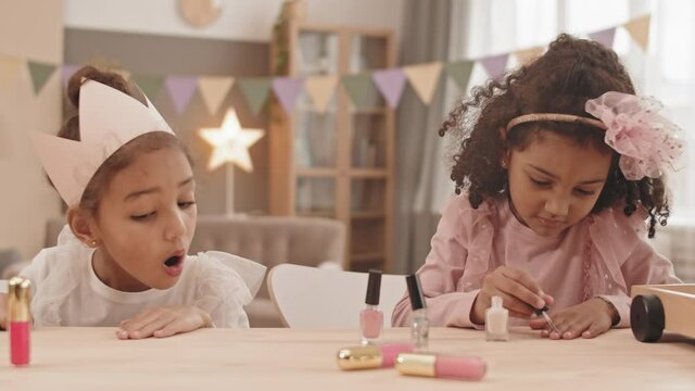 Chest-up of two cute Mixed-Race little girls sitting at table in living room, younger sister painting nails with polish, older child blowing on hands, drying product