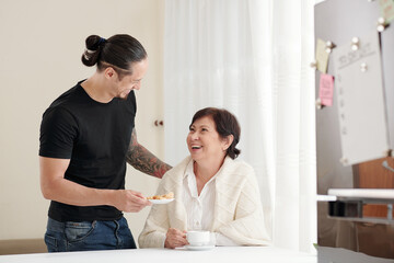 Cheerful caring adult son giving plate of homemade sugar cookies to mother visiting him at home