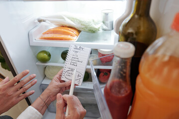 Hands of senior woman and her adult son writing shopping list when checking fridge