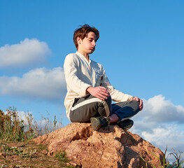 Naklejka na ściany i meble A Caucasian man from Spain sitting on a rock surrounded by grass and meditating on sky background