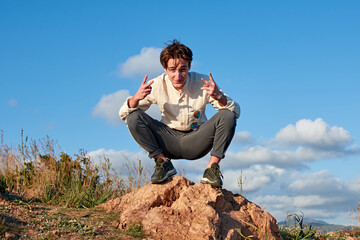 A Caucasian man from Spain making a peace sign with both hands while squatting on a rock surrounded...