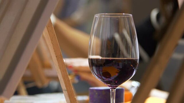A glass of wine at a painting master class. Female hands in the background paint a picture