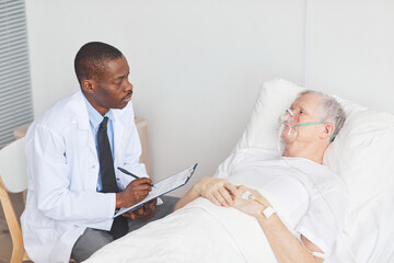 Obraz premium Portrait of African-American doctor talking to senior man lying in hospital bed with oxygen supplementation mask, copy space