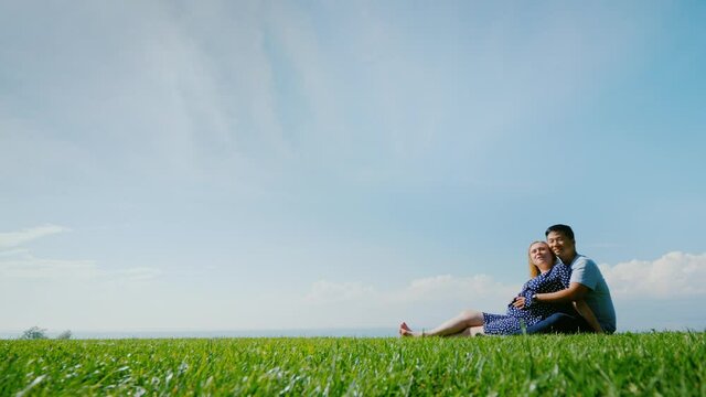 Asian man with a pregnant woman are resting in nature in a picturesque place, sit on the green grass
