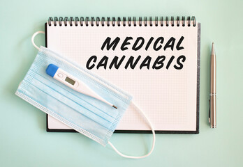 A medical protective mask and a thermometer are on the notebook. MEDICAL CANNABIS text in a notebook.