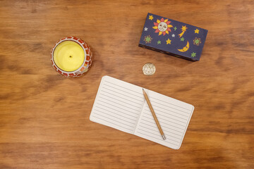 Still life of a pencil on an open notebook, candle, wooden box and medallion. Wood background.