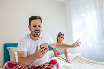 Confused young couple having an argument about mobile phone while lying in bed at home. Relationship problems, cheating online or great offer and good ad.