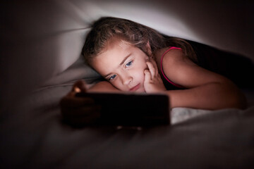 A girl is seriously looking on a smartphone at night under a blanket. Children's loneliness, dependence on gadgets.