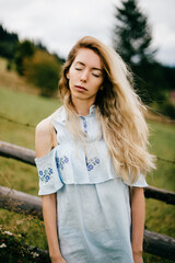 Young attractive elegant blonde girl in blue romantic dress posing near fence in the countryside