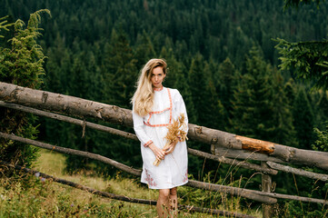 Young attractive blonde girl in white dress with embroidery posing with spikelets bouquet over...