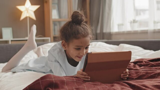Full shot of adorable Mixed-Race little girl wearing hair in bun, lying on stomach in bed in her room, using tablet computer