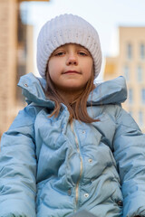 little girl portrait wearing white knitted hat and sports jacket have fun on outdoor playground during sunny cold spring vacation. Funny playful little kid outside high-rise buildings with apartments