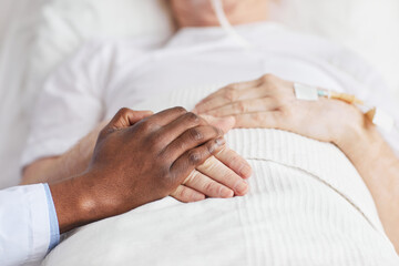 Close up of caring African-American doctor holding hands with senior patient lying in hospital bed, copy space