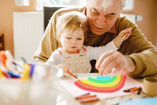 Cute little baby toddler girl and handsome senior grandfather painting with colorful pencils at home. Grandchild and man having fun together. Family and generation in love