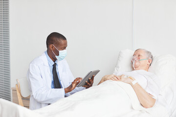 Portrait of African-American doctor wearing mask while talking to senior man lying in hospital bed, copy space