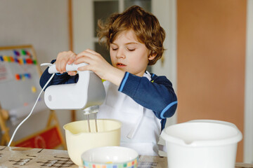 Adorable funny blond little kid boy baking chocolate cake and tasting dough in domestic kitchen, indoors. Happy child having fun with working with mixer, flour, eggs dough at home. Little helper