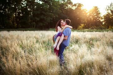 Naklejka na ściany i meble A man holds a woman in his arms in a field at sunset. A couple in love spend the evening walking together. A brunette in a long dress smiles and hugs her lover. Heterosexual couple enjoy each other