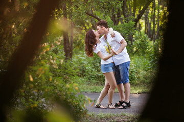 Naklejka na ściany i meble A young couple in love walks in a summer park at sunset and kisses and hugs hard. The heterosexual couple is wearing the same outfit, white T-shirts and shorts. Tender relationship between lovers