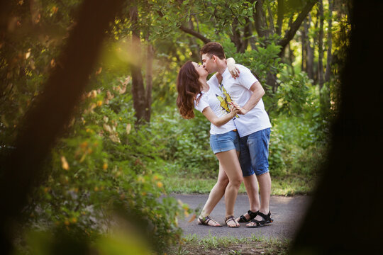 A young couple in love walks in a summer park at sunset and kisses and hugs hard. The heterosexual couple is wearing the same outfit, white T-shirts and shorts. Tender relationship between lovers