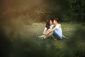 A guy and a girl are sitting on the grass and looking at each other in the park. A loving couple smiles and relaxes while sitting on the lawn. Heterosexual couple human love relationship concept