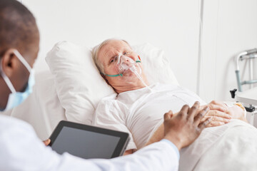 Portrait of sick senior man lying in hospital bed with oxygen mask and talking to African-American doctor comforting him, copy space