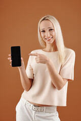 Naklejka premium Portrait of smiling young pretty woman pointing at screen of smartphone in her hand and looking at camera