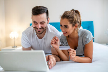 Lovely young happy couple laying in bed, shopping online with credit card and laptop computer. Beautiful young amorous couple using laptop, on bed. Happy young couple shopping online 