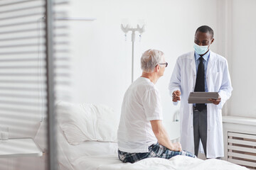 Portrait of African-American doctor talking to senior man in white hospital room, copy space