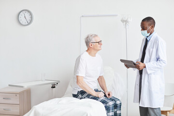 Obraz premium Portrait of recovering senior man talking to African-American doctor while sitting on bed in white hospital room, copy space