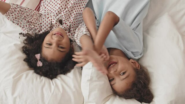 Top view medium close-up POV of two brown-eyed Mixed-Race girls lying in bed smiling, moving hands, looking on camera