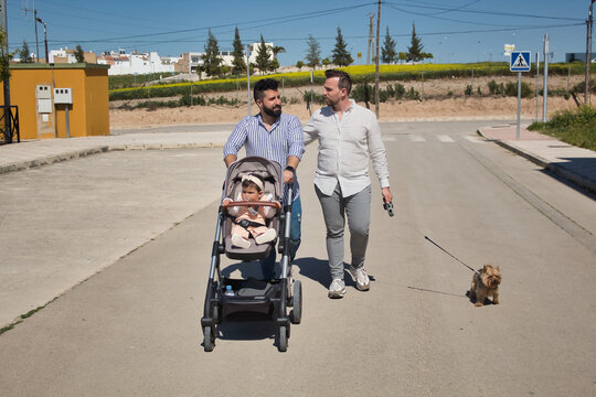 Gay Married Couple Walking Their Daughter In A Stroller In The Street With Their Dog.