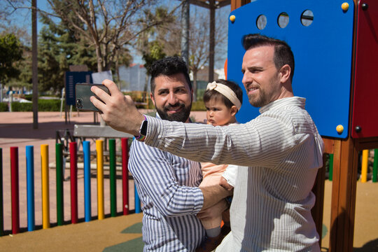 Gay married couple taking a selfie with their cell phone with their daughter in their arms in a playground.