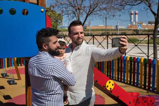 Gay Married Couple Taking A Selfie With Their Cell Phone With Their Daughter In Their Arms In A Playground.