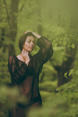 Beautiful young woman in a black transparent chiffon dress among the green foliage of forest trees