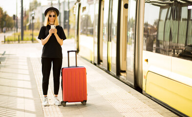 Traveler tourist woman in black casual clothes, sunglasses and hat with red suitcase use mobile phone to buy tickets online and sit on bus station. Female passenger travel abroad on weekends getaway