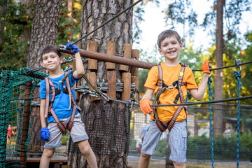 Two cute happy young children, boy and his brother in protective harness, carbine and safety helmets on rope way on bright sunny day on green foliage bokeh background.