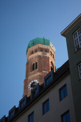 Fototapeta na wymiar München: Der Turm der Frauenkirche an einem sonnigen Tag mit blauem Himmel. Kontrast zwischen den gewöhnlichen Häusern im Vordergrund und der prächtigen Kirche im Hintergrund 