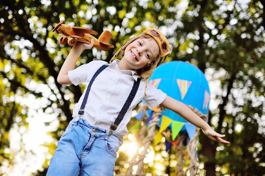 little boy in a white shirt with suspenders with curly hair in a pilot's hat and glasses plays with a wooden plane against the background of a basket with a blue balloon.