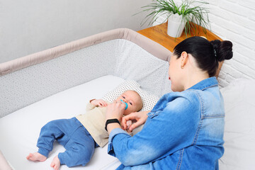 mother plays with a baby who is lying in a side-bed for a newborn or in a modern playpen