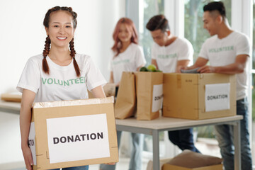 Cheerful pretty young Vietnamese woman holding big box of groceries packed for charity center or...
