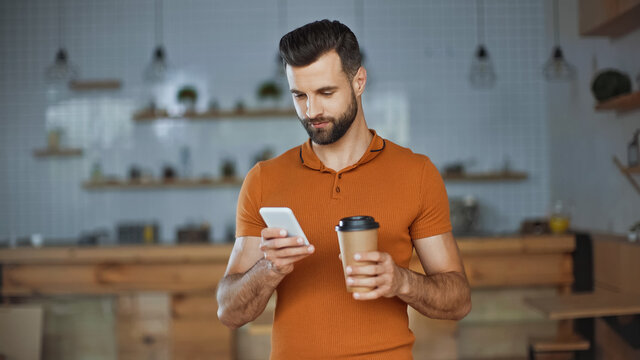 bearded man using smartphone and holding coffee to go in cafe