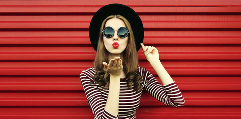 Portrait close up of beautiful young woman blowing her red lips sending air kiss wearing a black round hat on a background