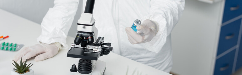 Cropped view of scientist in protective uniform holding vaccine near microscope and pills, banner