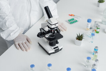 Cropped view of scientist in hazmat suit standing near microscope and vaccines on table
