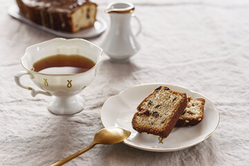 Slice of Pound Loaf Cake with raisins on white porcelain plate and cup of tea on greige linen tablecloth. Selective focus