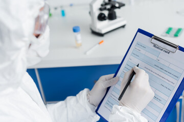 Fototapeta premium Medical card and pen in hands of scientist in protective uniform on blurred foreground
