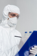 Scientist in hazmat suit and medical mask writing on clipboard on blurred foreground